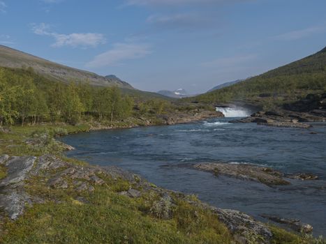 Beautiful landscape with waterfall on wild Tjaktjajakka river, birch tree forest and mountains. Lapland naturein summer at Kungsleden hiking trail. Blue sky background.