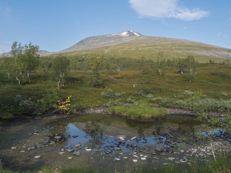 Colorful snow capped mountain Sanjartjakka reflecting in small pond and Beautiful wild Lapland nature landscape and birch tree forest. Summer blue sky background.