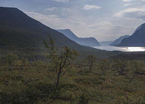 Beautiful wild Lapland nature landscape with blue Kaitumjaure lake, birch tree forest and mountains. Northern Sweden summer at Kungsleden hiking trail. Blue sky background.