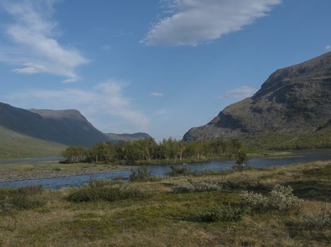 Confluence of wild Tjaktjajakka river and Kaitumjaure lake with small birch tree forest island and mountains. Lapland nature landscape in summer at Kungsleden hiking trail