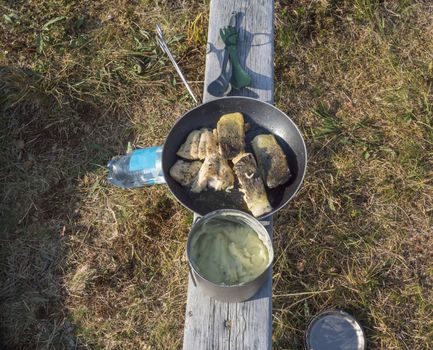 Freshly cooked mashed potatoes and fried trout fish fillets on cooking pan and mess tin on wooden board. Outdoor camping cooking