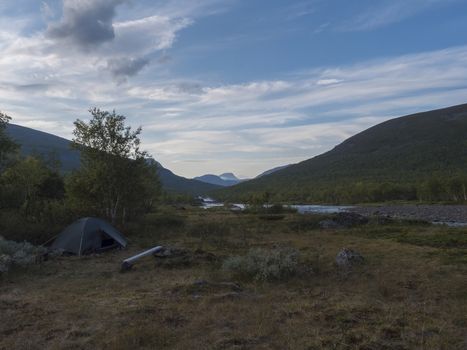 Small green tent in Beautiful wild Lapland nature landscape with blue river, Kaitumjaure lake, birch trees and mountains. Northern Sweden summer at Kungsleden hiking trail. Blue sky dramatic clouds.