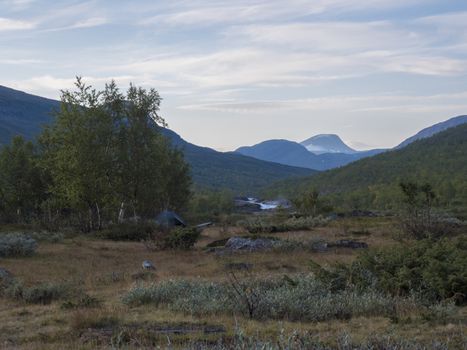 Beautiful landscape with small green tent, waterfall on wild Tjaktjajakka river, birch tree forest and mountains. Lapland naturein summer at Kungsleden hiking trail. Blue sky background.