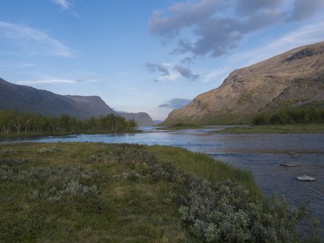 Confluence of wild Tjaktjajakka river and Kaitumjaure lake with small birch tree forest island and mountains. Lapland nature landscape in summer at Kungsleden hiking trail