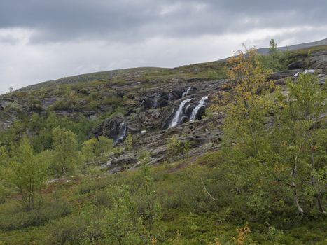 waterfall cascade, birch tree forest and mountains. Lapland nature landscape in summer, moody sky