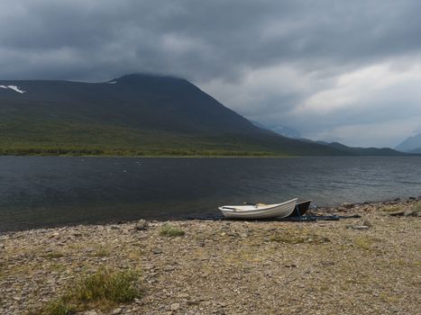 two rowing boats at Kungsleden hiking trail on bank of Teusajaure lake with birch tree forest and mountains. Lapland nature landscape in summer, moody sky
