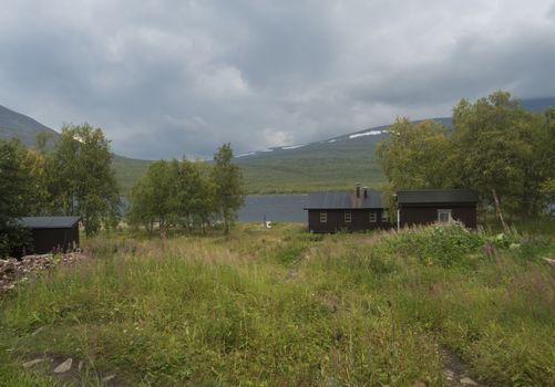 STF Teusajaure Fjallstuga Mountain cabin hut with blue Teusajaure lake, green hills, birch forest and footpath of Kungsleden hiking trail. Lapland nature landscape in summer, moody sky
