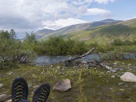 hiking boots with view on wild river with birch tree forest and mountains in golden light. Lapland nature landscape in summer, moody sky