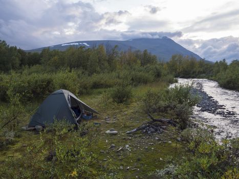 Small green tent in Beautiful wild Lapland nature landscape with blue river, birch trees and mountains. Northern Sweden summer at Kungsleden hiking trail. Blue sky dramatic clouds.