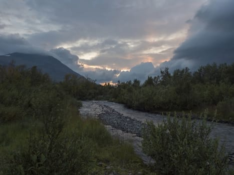 wild river with birch tree forest and mountains in sunset light. Lapland nature landscape in summer, moody sky, pink clouds