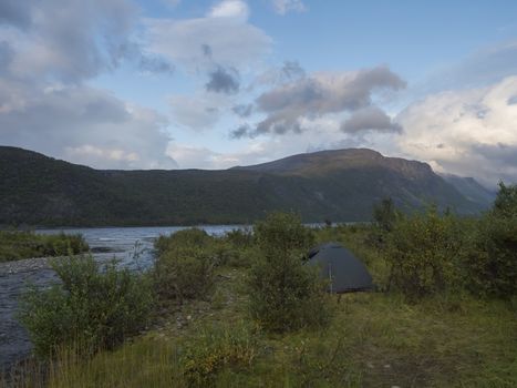 Small green tent in Beautiful wild Lapland nature landscape with blue river, birch trees and mountains. Northern Sweden summer at Kungsleden hiking trail. Blue sky dramatic clouds.