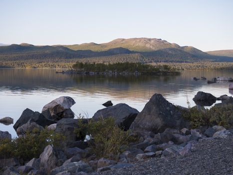 View on river Lulealven in Saltoluokta in Sweden Lapland during sunset golden hour. Green mountain, birch trees, rock boulders clouds and sky and clear the water