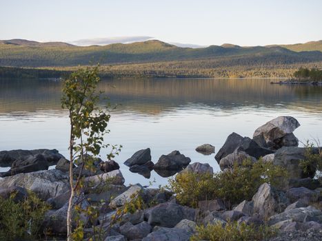 View on river Lulealven in Saltoluokta in Sweden Lapland during sunset golden hour. Green mountain, birch trees, rock boulders clouds and sky and clear the water