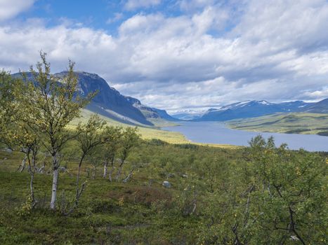 Lapland landscape with beautiful river Lulealven, snow capped mountain, birch tree and footpath of Kungsleden hiking trail near Saltoluokta, north of Sweden wild nature. Summer blue sky.