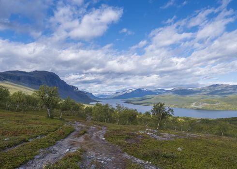 Lapland landscape with beautiful river Lulealven, snow capped mountain, birch tree and footpath of Kungsleden hiking trail near Saltoluokta, north of Sweden wild nature. Summer blue sky.