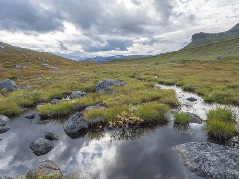Lapland landscape with small pond, stones boulders, autumn colored bushes, grass and snow capped mountain at Kungsleden hiking trail near Saltoluokta, Sweden. Blue sky white clouds