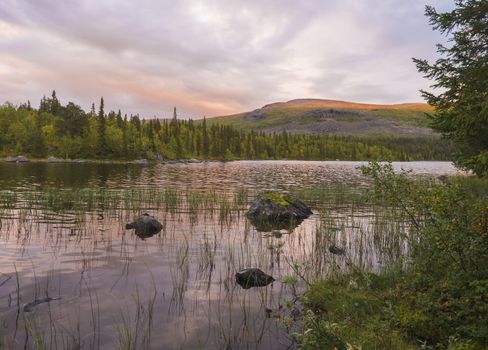 Orange pink sunset over lake Sjabatjakjaure in Parte in Sweden Lapland. Mountains, birch trees, spruce forest, rock boulders and grass. Sky, clouds and clear water