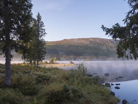 Beautiful morning sunrise over lake Sjabatjakjaure with haze mist in Sweden Lapland nature. Mountains, birch trees, spruce forest, rock boulders and grass. Sky, clouds and clear water