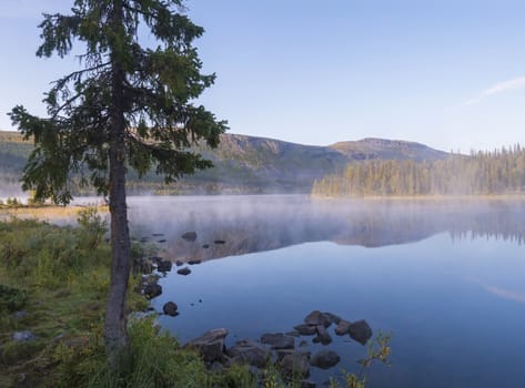 Beautiful morning sunrise over lake Sjabatjakjaure with haze mist in Sweden Lapland nature. Mountains, birch trees, spruce forest, rock boulders and grass. Sky, clouds and clear water