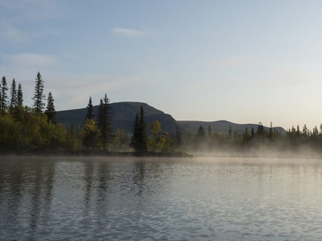 Beautiful morning sunrise over lake Sjabatjakjaure with haze mist in Sweden Lapland nature. Mountains, birch trees, spruce forest, rock boulders and grass. Sky, clouds and clear water