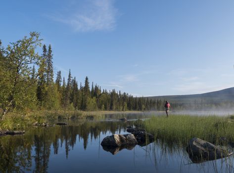 Fisherman man catching fish at lake Sjabatjakjaure in Beautiful sunny morning haze mist in Sweden Lapland nature. Mountains, birch trees, spruce forest. Blu sky.