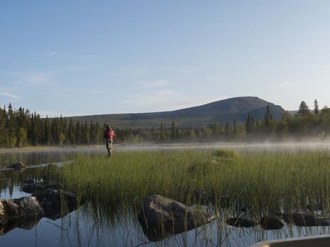 Fisherman man catching fish at lake Sjabatjakjaure in Beautiful sunny morning haze mist in Sweden Lapland nature. Mountains, birch trees, spruce forest. Blu sky.