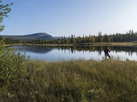 Fisherman man catching fish at lake Sjabatjakjaure in Beautiful sunny morning haze mist in Sweden Lapland nature. Mountains, birch trees, spruce forest. Blu sky.