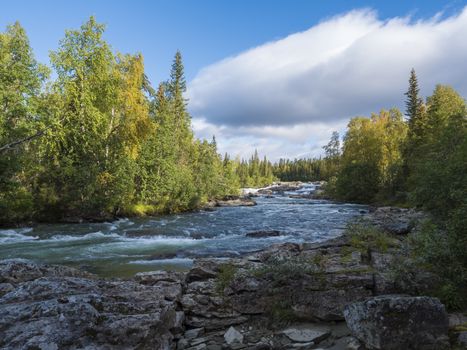 Fisherman man catching fish at beautiful northern landscape with azure blue river Kamajokk, mountains, boulders and spruce tree forest in Kvikkjokk in Swedish Lapland. Summer sunny day, white clouds.
