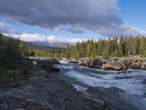Beautiful northern landscape with wild glacier river Kamajokk, boulders and spruce tree forest in Kvikkjokk in Swedish Lapland. Summer sunny day, golden hour, dramatic clouds.
