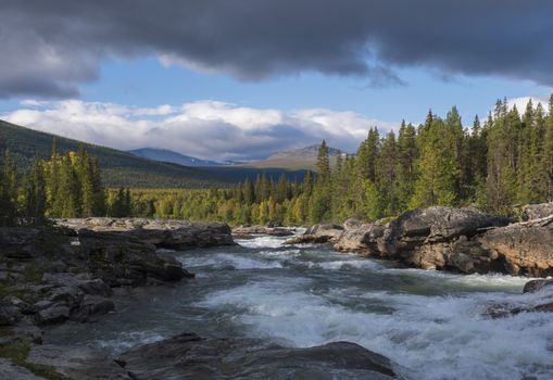 Beautiful northern landscape with wild glacier river Kamajokk, boulders and spruce tree forest in Kvikkjokk in Swedish Lapland. Summer sunny day, golden hour, dramatic clouds.