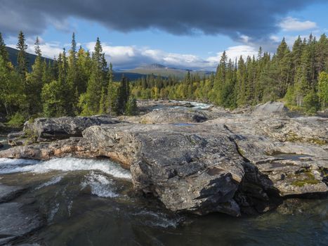 Beautiful northern landscape with wild glacier river Kamajokk, boulders and spruce tree forest in Kvikkjokk in Swedish Lapland. Summer sunny day, golden hour, dramatic clouds.