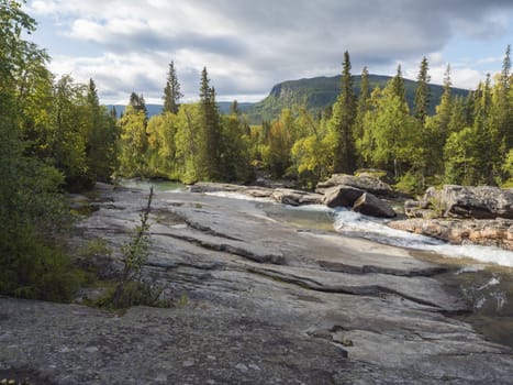Beautiful northern landscape with wild glacier river Kamajokk, boulders and spruce tree forest in Kvikkjokk in Swedish Lapland. Summer sunny day, golden hour, dramatic clouds.