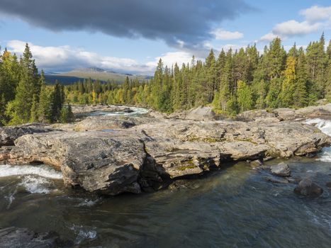 Beautiful northern landscape with wild glacial river Kamajokk, boulders and spruce tree forest in Kvikkjokk in Swedish Lapland. Summer sunny day, golden hour, dramatic clouds.
