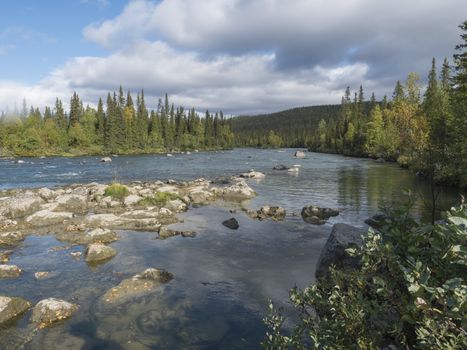 Beautiful northern landscape with wild glacier river Kamajokk, boulders and spruce tree forest in Kvikkjokk in Swedish Lapland. Summer sunny day, golden hour, dramatic clouds.
