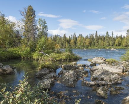 Beautiful northern landscape with wild glacial river Kamajokk, boulders and spruce tree forest in Kvikkjokk in Swedish Lapland. Summer sunny day, golden hour, dramatic clouds.