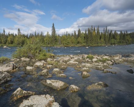 Beautiful northern landscape with azure blue river Kamajokk, mountains, boulders and spruce tree forest in Kvikkjokk in Swedish Lapland. Summer sunny day, white clouds.