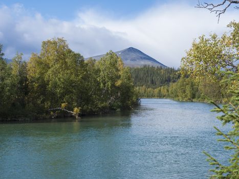 Beautiful northern landscape with azure blue river Kamajokk, mountains, boulders and spruce tree forest in Kvikkjokk in Swedish Lapland. Summer sunny day, white clouds.