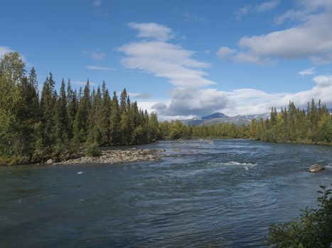 Beautiful northern landscape with wild glacial river Kamajokk, boulders and spruce tree forest in Kvikkjokk in Swedish Lapland. Summer sunny day, golden hour, dramatic clouds.