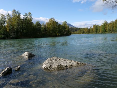 Beautiful northern landscape with azure blue river Kamajokk, mountains, boulders and spruce tree forest in Kvikkjokk in Swedish Lapland. Summer sunny day, white clouds.