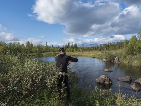 Fisherman man catching fish at beautiful northern landscape with azure blue river Kamajokk, mountains, boulders and spruce tree forest in Kvikkjokk in Swedish Lapland. Summer sunny day, white clouds.