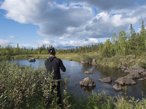 Fisherman man catching fish at beautiful northern landscape with azure blue river Kamajokk, mountains, boulders and spruce tree forest in Kvikkjokk in Swedish Lapland. Summer sunny day, white clouds.