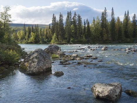 Beautiful northern landscape with azure blue river Kamajokk, mountains, boulders and spruce tree forest in Kvikkjokk in Swedish Lapland. Summer sunny day, white clouds.