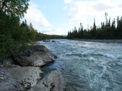 Beautiful northern landscape with wild glacial river Kamajokk, boulders and spruce tree forest in Kvikkjokk in Swedish Lapland. Summer sunny day, golden hour, dramatic clouds.