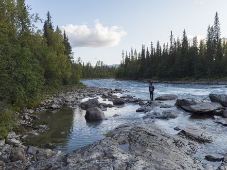 Beautiful northern landscape with wild glacial river Kamajokk, boulders and spruce tree forest in Kvikkjokk in Swedish Lapland. Summer sunny day, golden hour, dramatic clouds.
