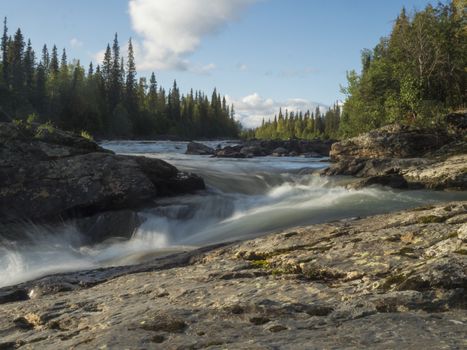 Beautiful northern landscape with long exposure water stream of river Kamajokk, boulders and spruce tree forest in Kvikkjokk in Swedish Lapland. Summer sunny day, golden hour, dramatic clouds.