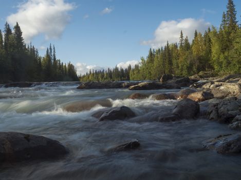 Beautiful northern landscape with long exposure water stream of river Kamajokk, boulders and spruce tree forest in Kvikkjokk in Swedish Lapland. Summer sunny day, golden hour, dramatic clouds.