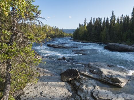 Fisherman man catching fish at beautiful northern landscape with azure blue river Kamajokk, mountains, boulders and spruce tree forest in Kvikkjokk in Swedish Lapland. Summer sunny day, white clouds.