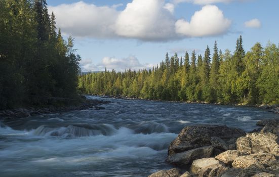 Beautiful northern landscape with long exposure water stream of river Kamajokk, boulders and spruce tree forest in Kvikkjokk in Swedish Lapland. Summer sunny day, golden hour, dramatic clouds.