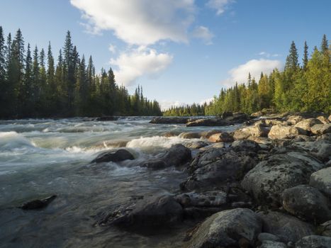 Beautiful northern landscape with long exposure water stream of river Kamajokk, boulders and spruce tree forest in Kvikkjokk in Swedish Lapland. Summer sunny day, golden hour, dramatic clouds.