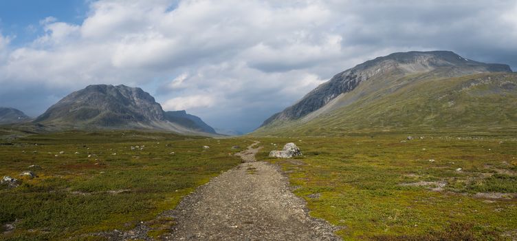 Wide panorama of Kungsleden hiking trail path with red marked stones in beautiful wild Lapland nature landscape with green bushes and mountains.Northern Sweden, summer day, blue sky, white clouds.